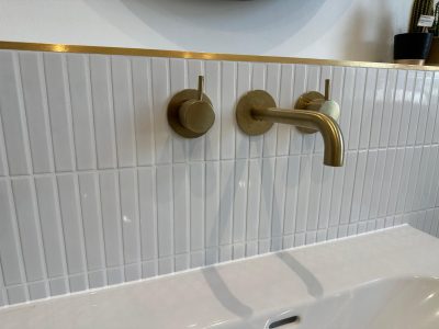 Brushed brass wall-mounted basin taps on white vertical mosaic tiles in South London bathroom