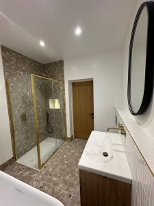 Wide angle view of shower enclosure with terrazzo tiles, brass frame, oak vanity and round mirror in South London bathroom