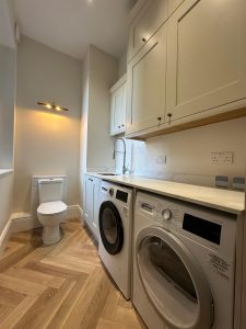 Combined utility room and WC with cream shaker cabinets, integrated washing machines and herringbone floor in South London