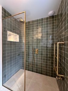 Walk-in shower enclosure with dark green square tiles, brass frame and brass ladder towel rail in South London